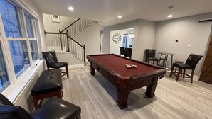A pool table in a room with a staircase, chairs, and a clock on the wall.