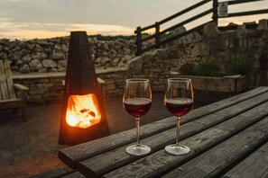 Two wine glasses on a wooden bench with a stone wall and a fire pit in the background.