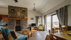 A cozy living room with a stone fireplace, a television, and a dining area with a table and chairs. - The Rock, Creevy Cottages (Rossnowlagh)