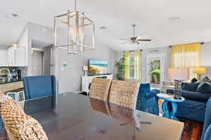 A modern living room with a glass dining table, blue chairs, a sofa, a ceiling fan, and a view of the beach through a door.