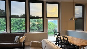 A living room with a sofa, a dining table with chairs, and large windows with a view of trees and hills. - Riverside Loft with Huge Deck on GAP Trail (Confluence)
