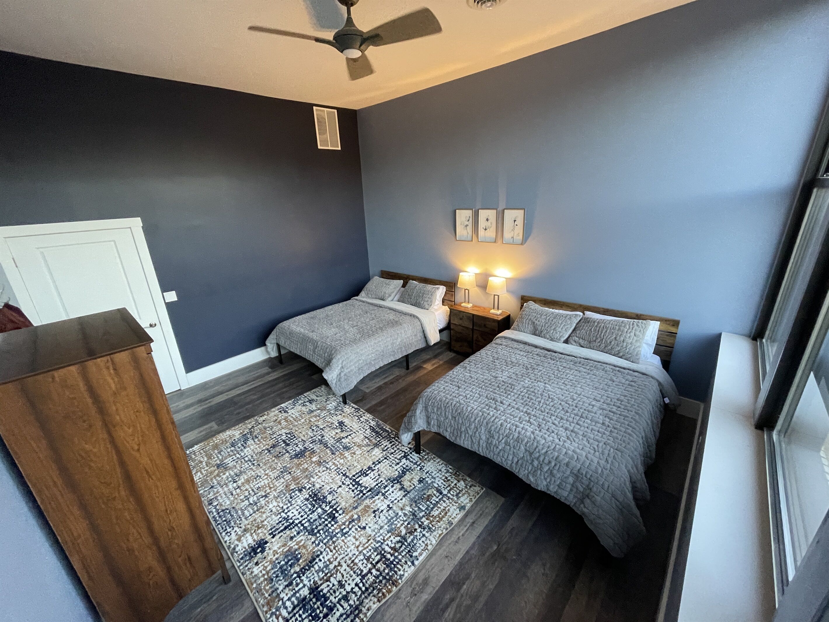 A bedroom with two beds, a ceiling fan, a nightstand with lamps, and a patterned rug.