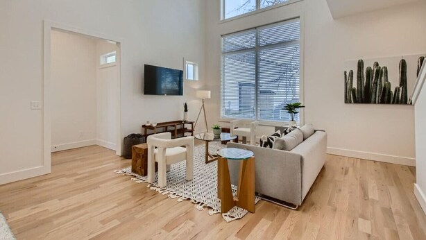 Ein großzügiger Wohnraum mit hohen Decken, einem großen Fenster mit Jalousien, einem an der Wand befestigten Flachbildfernseher, einem grauen Sofa, einem Couchtisch und einem gemusterten Teppich.