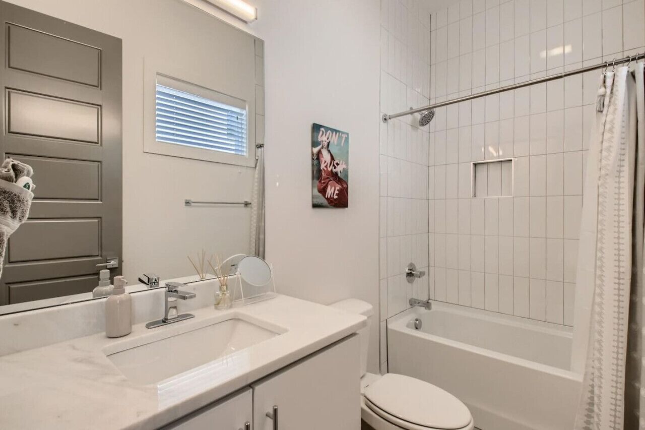 Une salle de bain avec un lavabo blanc, un miroir et des toilettes. On y trouve une douche avec une porte en verre et un porte-serviettes. Un poster est accroché au mur, et une fenêtre avec des stores est visible.
