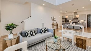 Ein modernes Wohnzimmer mit einem grauen Sofa, einem Glastisch und einem Essbereich mit weißem Tisch und Stühlen.