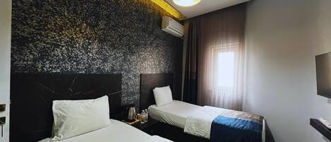 Habitación de hotel con dos camas, cabecero moderno, luz de techo, unidad de aire acondicionado y ventana con cortinas.