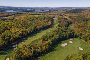 An aerial view of a golf course surrounded by dense forest with multiple water bodies in the background.