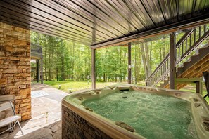 A hot tub with a stone wall and a wooden deck, surrounded by a forested area. - Relax, Play w/ Lake Days & Fire Pit Nights (Swanton)