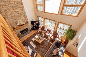 A spacious living room with a stone fireplace, a flat-screen TV, and a large window overlooking a forest. - Relax, Play w/ Lake Days & Fire Pit Nights (Swanton)