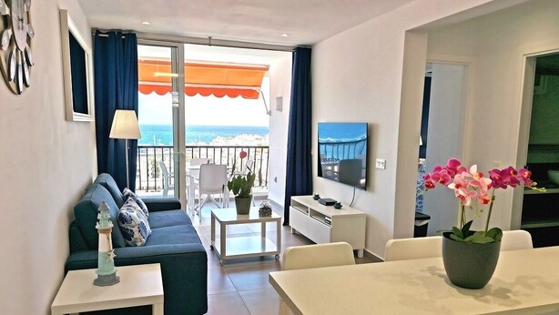 Sala moderna com sofá, mesa de jantar e televisão. Varanda com vista para o mar.