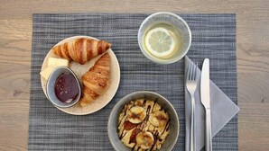 A breakfast setting with a croissant, a bowl of oatmeal with banana slices, a cup of tea, and a small bowl of jam on a textured placemat. - Boje06 (Heringsdorf)
