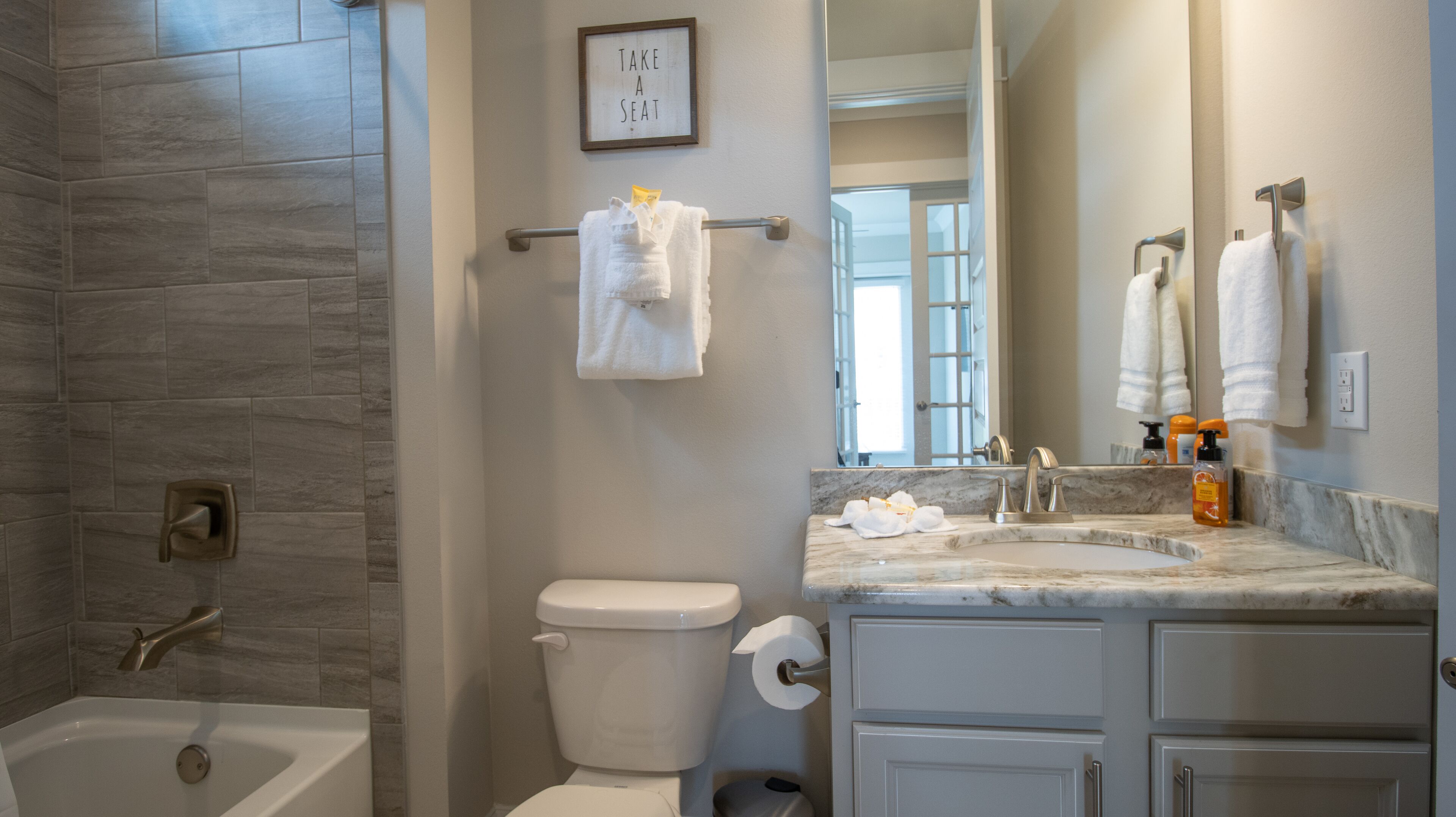 Une salle de bain avec une baignoire, des toilettes et un lavabo. On y trouve un porte-serviettes avec des serviettes et un panneau encadré sur lequel est écrit : « ASSEYEZ-VOUS ».