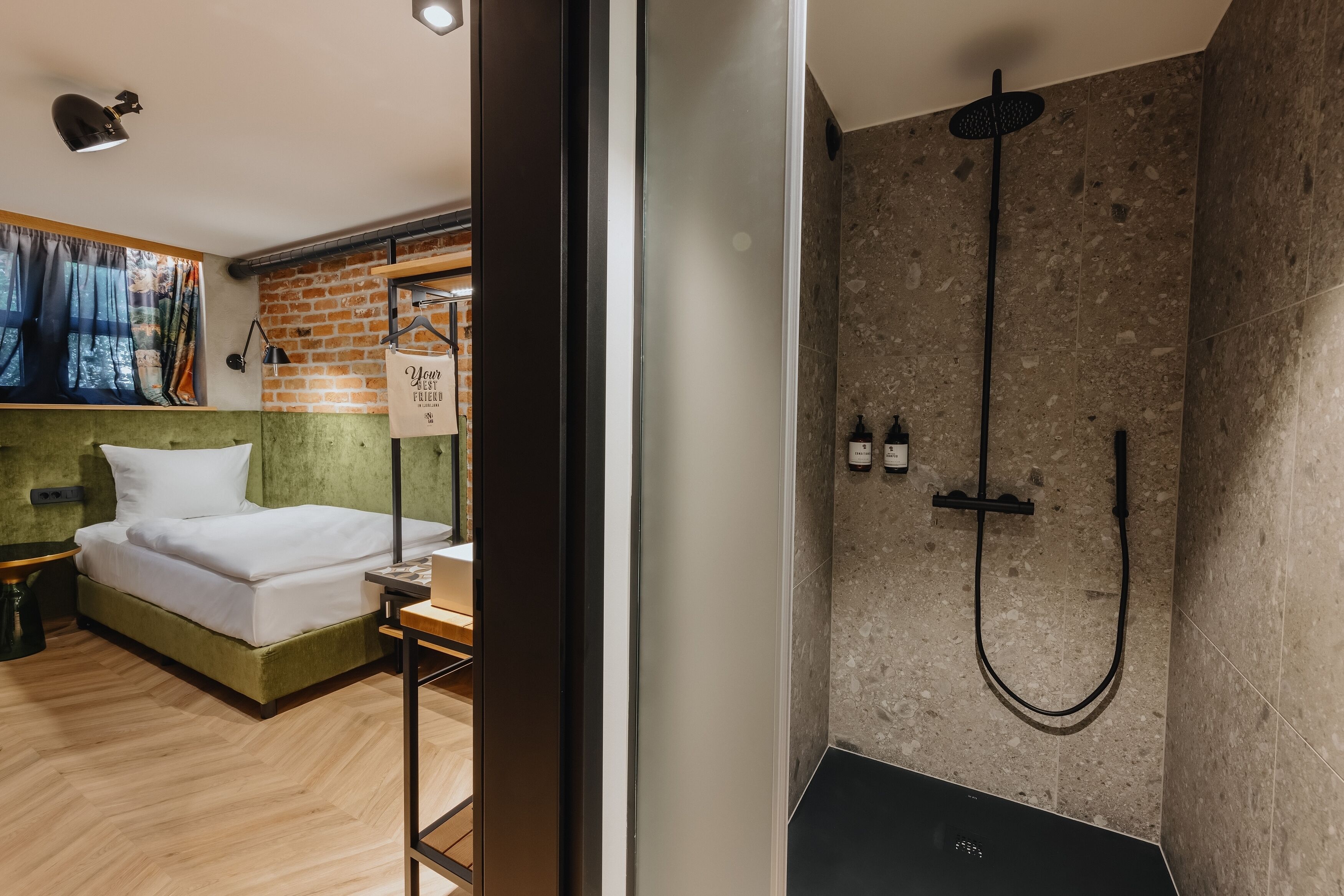 A modern hotel room with a bed, a small table, a nightstand, a brick accent wall, a shower with a handheld showerhead, and a window with curtains.