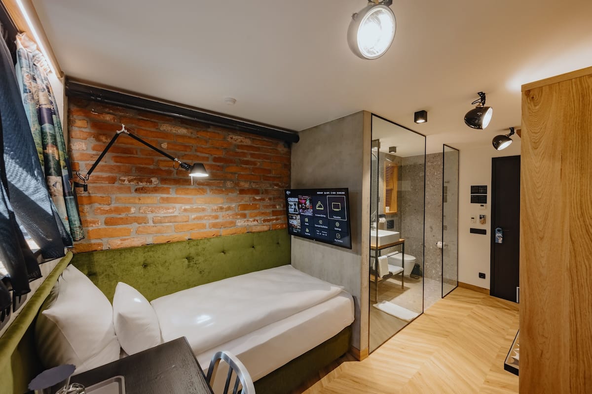 Quarto moderno com parede de tijolos, cabeceira estofada verde, cama, escrivaninha com computador e box de vidro.