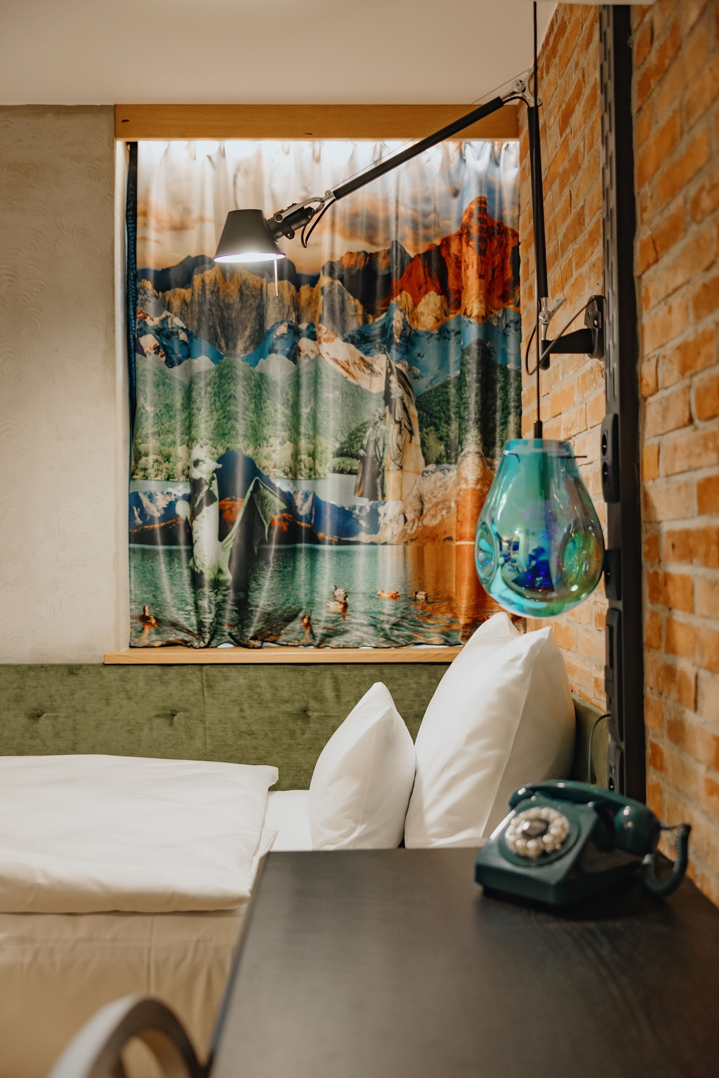Una habitación con cama, una mesa con teléfono, una lámpara colgante y una ventana con cortina que muestra un paisaje de montaña.