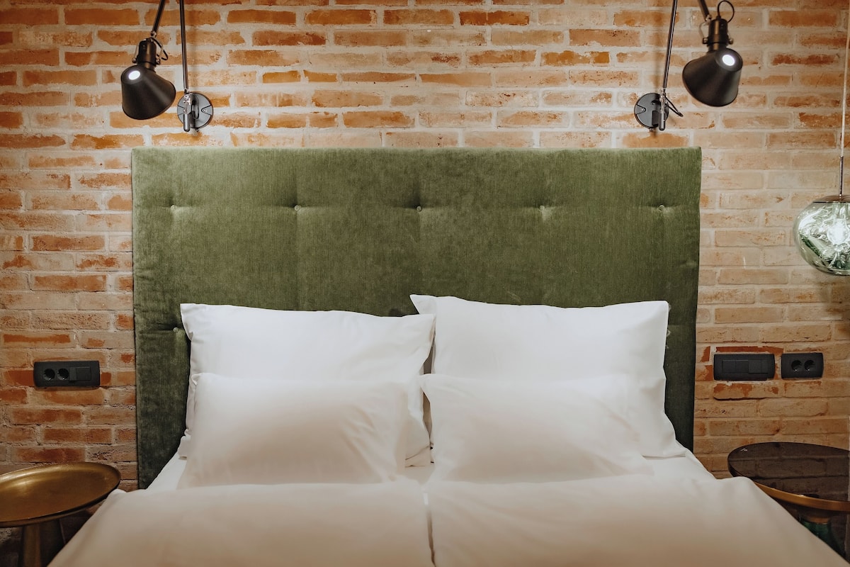 Uma cama com cabeceira estofada verde, almofadas brancas e duas luminárias pretas fixadas na parede.