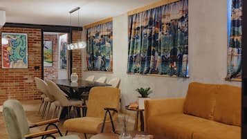 A modern living room with a brick wall, a dining area with a round table and chairs, and large windows with curtains.