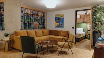 Ett modernt vardagsrum med en hörnsoffa, ett soffbord och ett litet sidobord. Det finns ett stort fönster med gardiner, en affisch på väggen och en dörr som leder till ett sovrum.