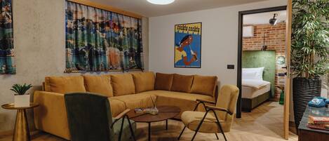 A modern living room with a sectional sofa, a coffee table, and a small side table. There is a large window with curtains, a poster on the wall, and a door leading to a bedroom.