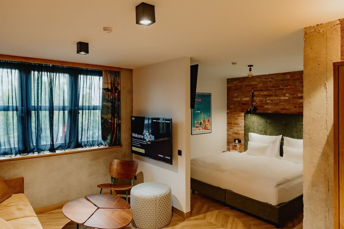 Quarto de hotel moderno com uma cama grande, televisão, sofá e área de jantar com padrão herringbone no chão.