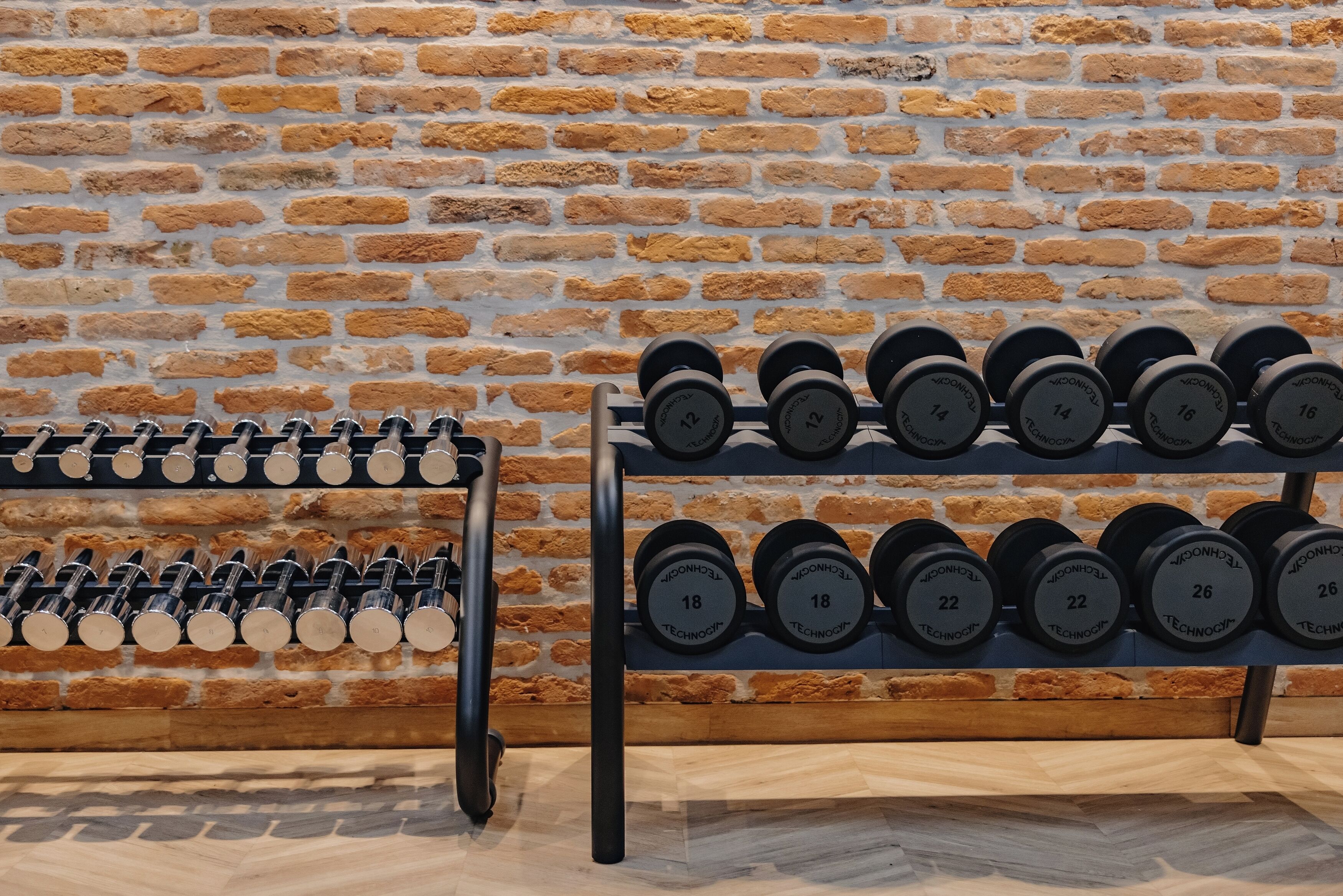 A wall of dumbbells with visible weight markings, arranged on shelves against a brick wall.