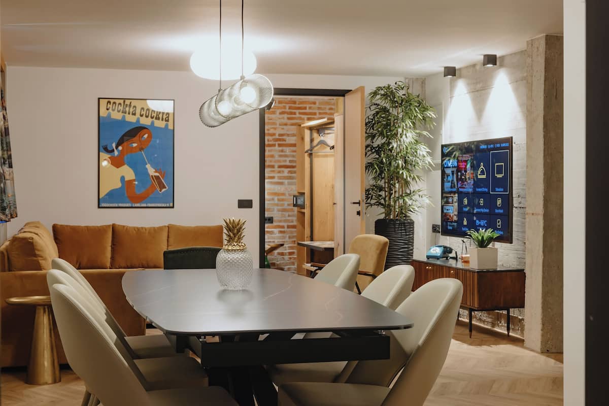 Uma sala de estar moderna com uma mesa de jantar, sofá e cadeiras. Há uma planta grande em vaso, uma TV montada na parede e um pôster.