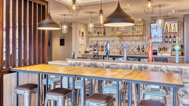 Een bar met een lange houten toonbank, metalen krukken, hangende吊り下げ型ライト en een bar gevuld met flessen.