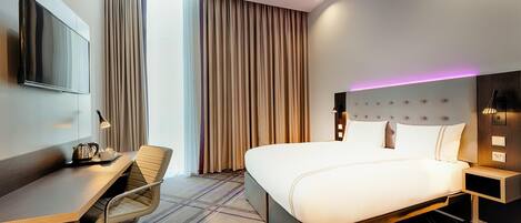 A hotel room with a bed, a desk, a chair, a television, a nightstand, and a window with curtains.