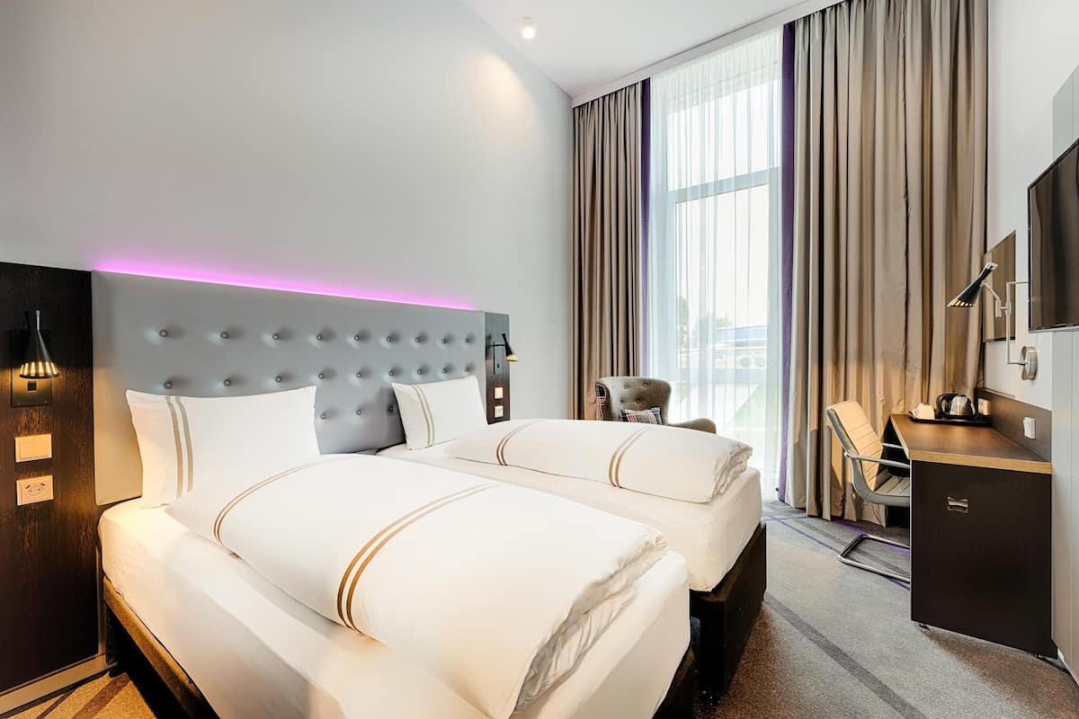 A hotel room with two beds, a desk, a chair, a TV, and a large window with curtains.