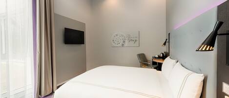 A modern hotel room with a large bed, a flat-screen TV mounted on the wall, a small table with a chair, and a decorative wall clock.