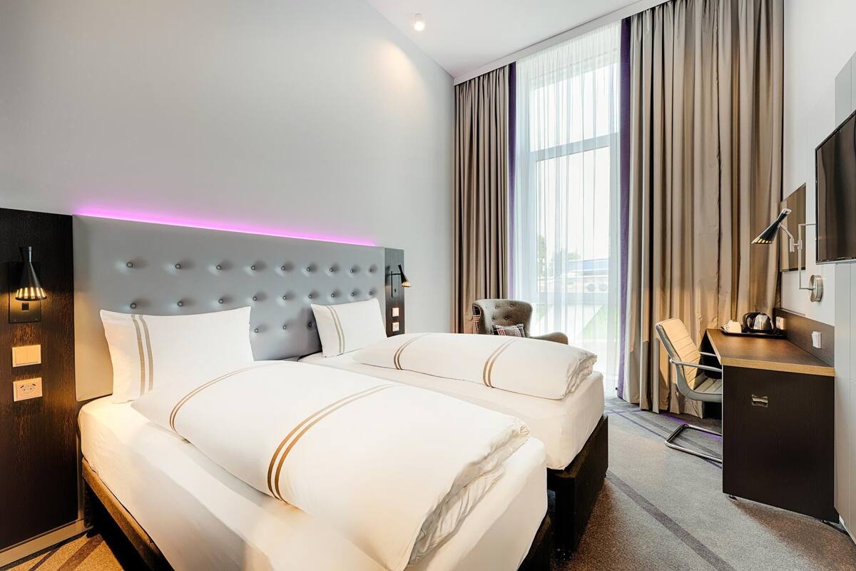 Ein Hotelzimmer mit zwei Betten, einem Schreibtisch, einem Stuhl, einem Fernseher und einem großen Fenster mit Vorhängen.