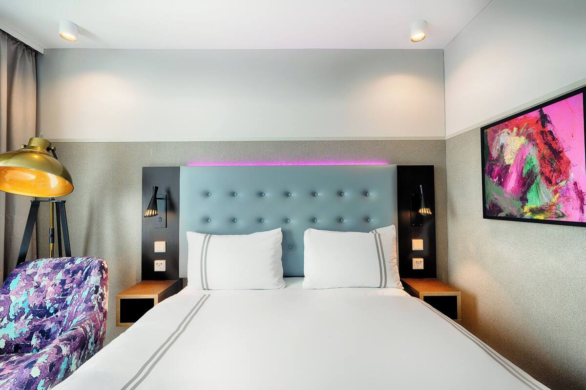 Ein modernes Hotelzimmer mit einer geblümten Kopfteil, einem großen Bett, einem farbenfrohen Sessel, Nachttischlampen und einem lebendigen, abstrakten Gemälde an der Wand.
