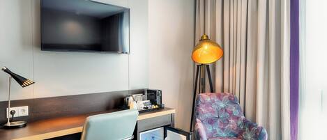 Habitación de hotel con cama, escritorio con silla, televisor montado en la pared, refrigerador pequeño, lámpara y ventana con cortinas.
