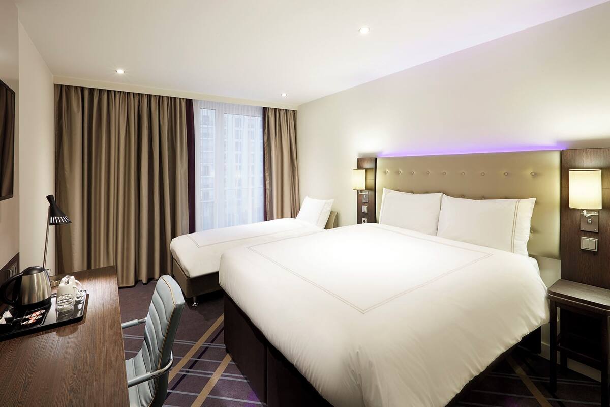 Ein Hotelzimmer mit zwei Betten, einem Schreibtisch mit einem Tablett mit Annehmlichkeiten, einem Stuhl und einem großen Fenster mit Vorhängen.