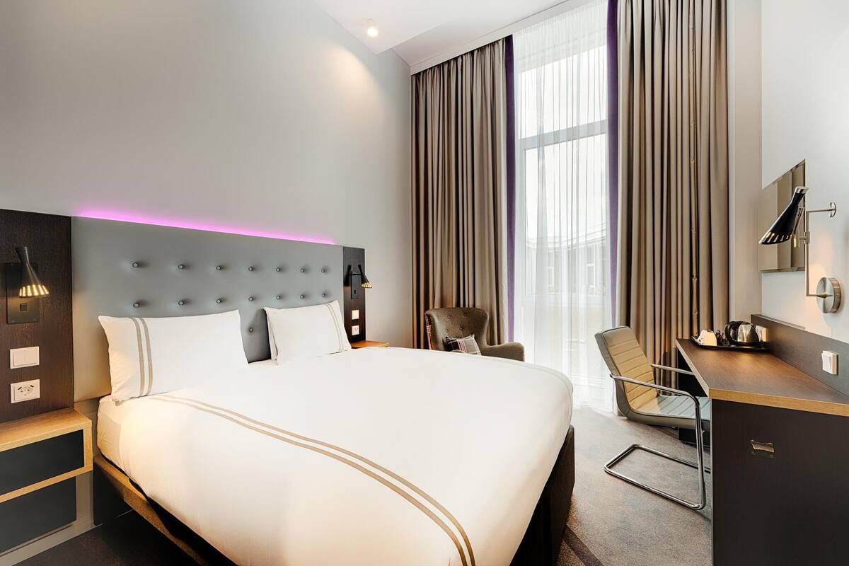 Ein modernes Hotelzimmer mit einem großen Bett, einem Schreibtisch, einem Sessel und einem großen Fenster mit Vorhängen.