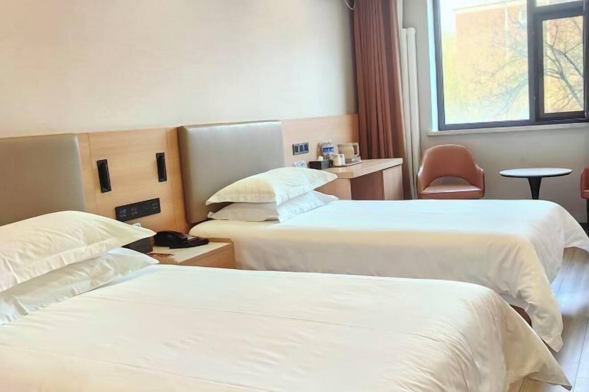 A hotel room with two beds, a bedside table with a lamp, a chair, and a window with curtains.