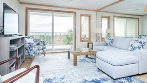A spacious living room with a large window, a ceiling fan, a sofa, a coffee table, and a TV on a stand. - Upgraded Oceanfront 3BR Condo in WrightsvilleDunes (Wrightsville Beach)