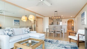 A modern living room with a light blue sofa, a glass coffee table, and a dining area with a wooden table and chairs. - Upgraded Oceanfront 3BR Condo in WrightsvilleDunes (Wrightsville Beach)