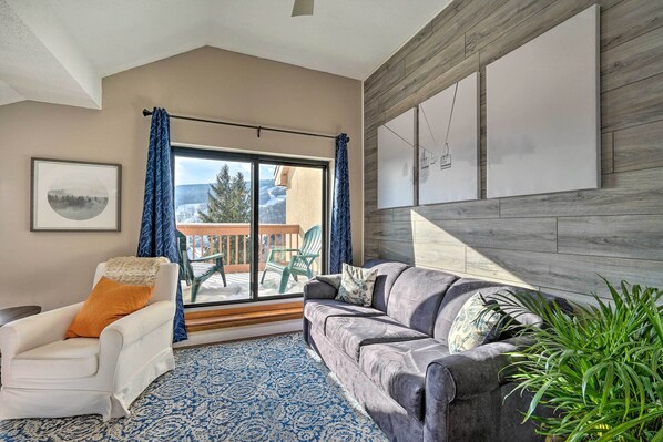 Una sala de estar con un sillón blanco, un sofá gris, un ventanal con cortinas, un tapete azul estampado y una planta verde.