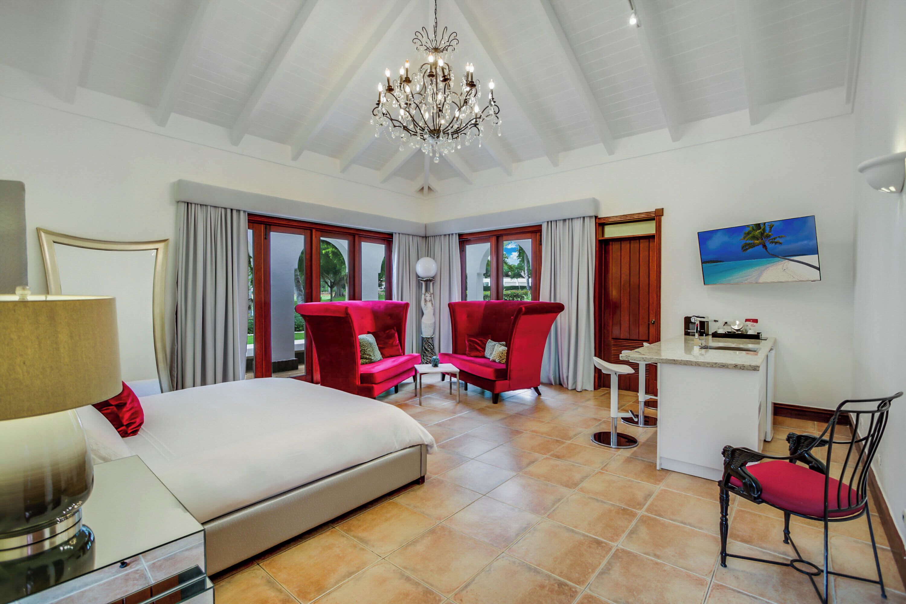 A bedroom with a bed, a desk, two red chairs, a chandelier, and a painting of a beach.