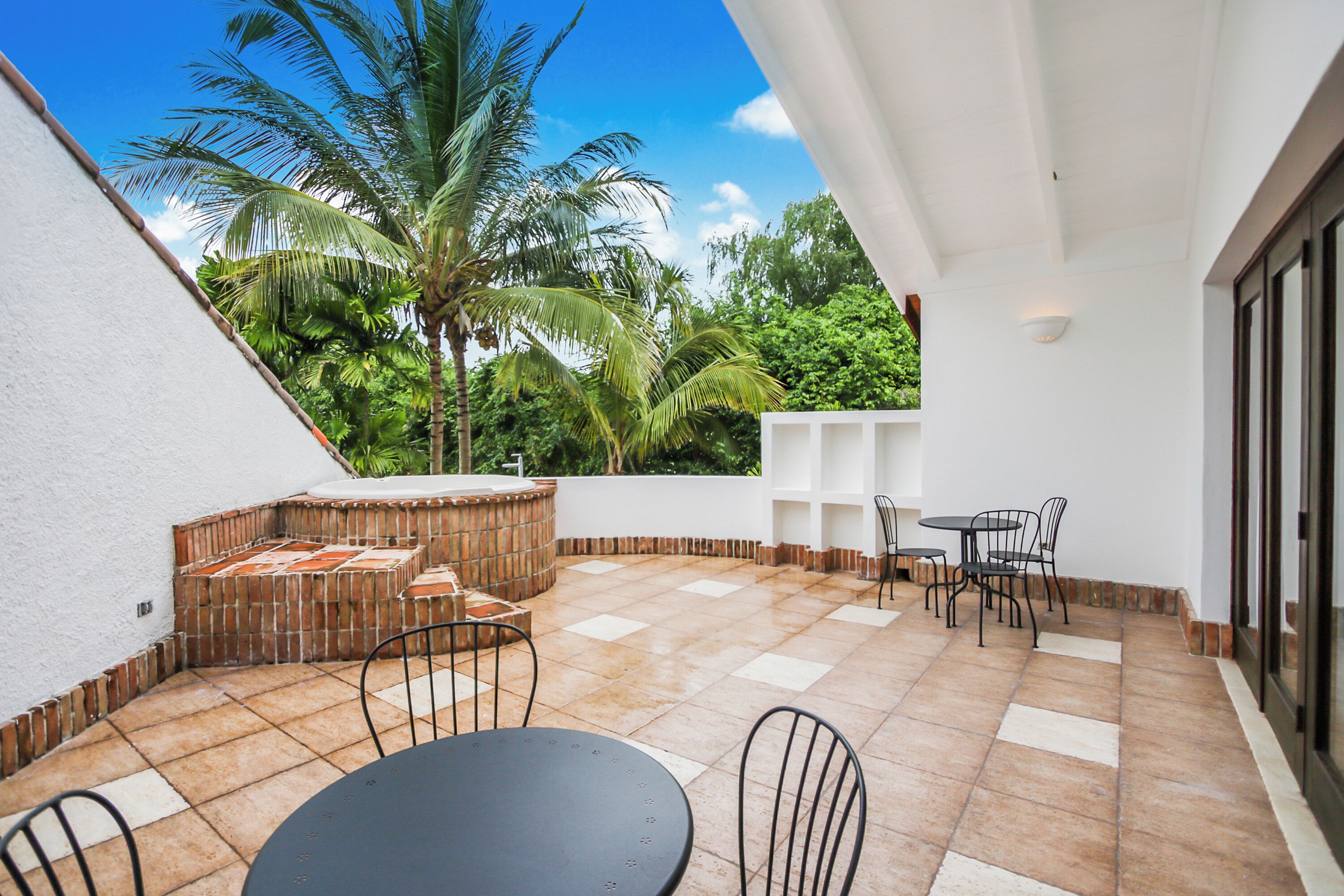 A patio with a round table and chairs, a small seating area with cushions, and a view of palm trees and greenery.