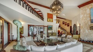 A spacious living room with a large white sofa, a glass coffee table, and a chandelier. There is a staircase with a wooden railing leading to an upper level. - 5-star villa for rent in Moroccan-style (La Romana)