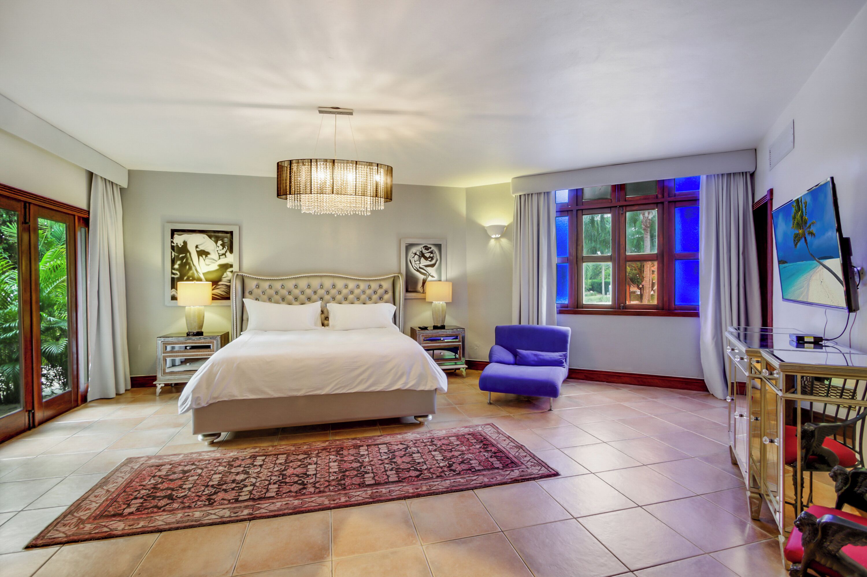 A spacious bedroom with a large bed, a chandelier, a blue armchair, and a television on a stand.