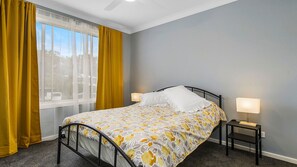 Ein Schlafzimmer mit einem Bett, zwei Nachttischen, einem Fenster mit gelben Vorhängen und einem Deckenventilator.