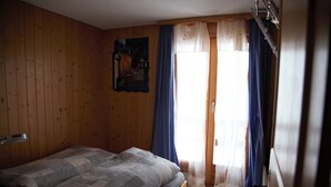A bedroom with a wooden wall, a bed with a grey and white checkered bedspread, a painting on the wall, and a window with blue curtains. - This stunning, bright and comfortable chalet can accommodate up to 8 people. Located on the Chemin d (Les Masses)