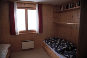 Un dormitori amb parets de fusta, una finestra amb cortines i un llit amb llençols estampats florals, a més d'una prestatgeria.