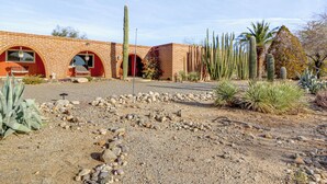 Um edifício em estilo desértico com paredes de tijolo e janelas em arco, rodeado por diversos cactos e plantas típicas do deserto.