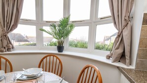 A dining area with a round table set for four, surrounded by wooden chairs. A potted plant is on the windowsill, and the room has a view of the outside.