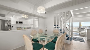 Et moderne køkken med hvide skabe, et rustfrit stål køleskab og et spisebord med glasplade omgivet af fletstole med grønne hynder.