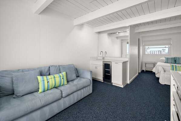 A modern, well-lit room with a grey sofa, a small kitchenette, and a window providing a view of the cityscape. - Ocean Colony-Enjoy Ocean Views at this beautiful Condo! (Ocean City)